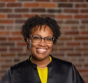 Black women in front of brick wall, smiling, wearing yellow shirt and black dress coat.